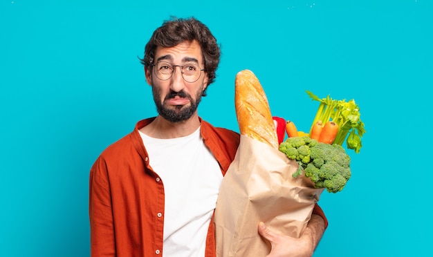 Young bearded man feeling sad and whiney with an unhappy look, crying with a negative and frustrated attitude and holding a vegetables bag