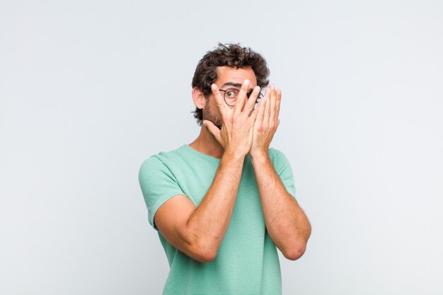 Young bearded man feeling scared or embarrassed, peeking or spying with eyes half-covered with hands