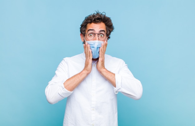 Young bearded man feeling shocked and scared, looking terrified with open mouth and hands on cheeks