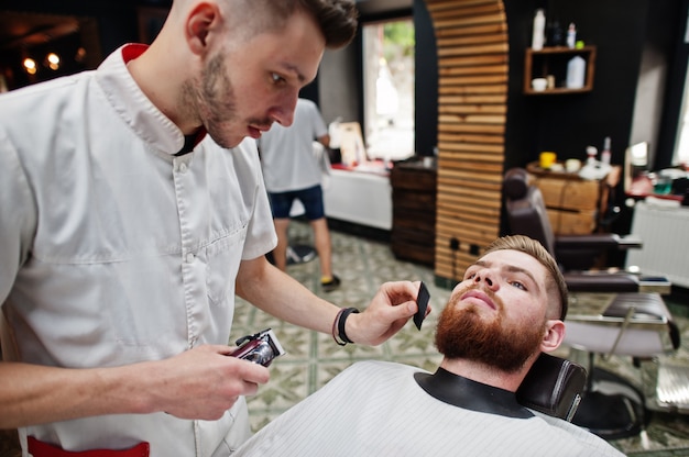 Young bearded man getting haircut by hairdresser while sitting in chair at barbershop. Barber soul.