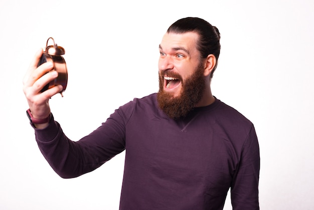 young bearded man is looking stressed at a clock that he is holding near a white wall