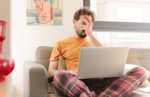 Young bearded man looking shocked scared or terrified covering face with hand and peeking between fingers and sitting with a laptop