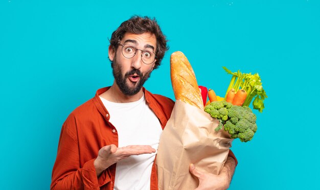 Young bearded man looking surprised and shocked, with jaw dropped holding an object with an open hand on the side and holding a vegetables bag