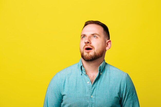 Young bearded man looks amazed funny male dressed in blue shirt is funny studio portrait on yellow b...