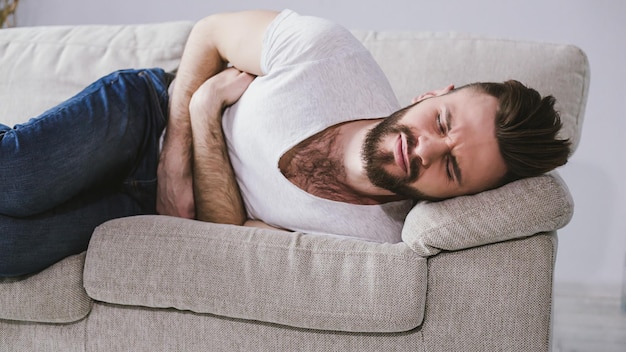 Young bearded man lying on a couch holding onto his stomach because of severe pain