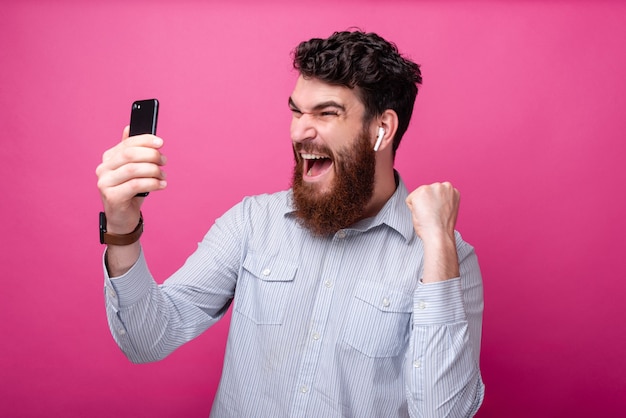 Young bearded man making a winner gesture with one hand while looking at his phone, yelling and wearing earpods.