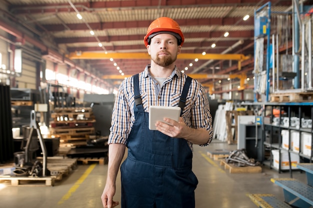 Young bearded man in overalls and helmet posing in one of large factory workshops