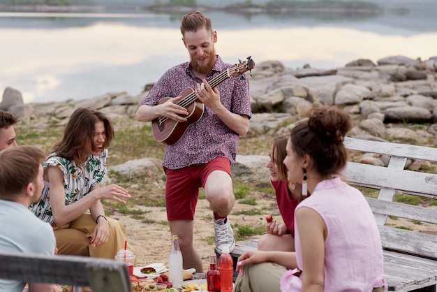 Young bearded man playing the guitar and smiling joyfully while hanging out with friends near lake, sitting around table, eating and talking