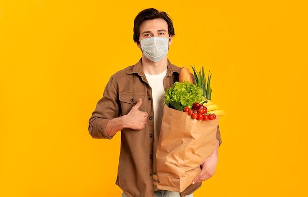 Young bearded man in protective medical mask with paper package of vegetables products shows thumbs up gesture isolated