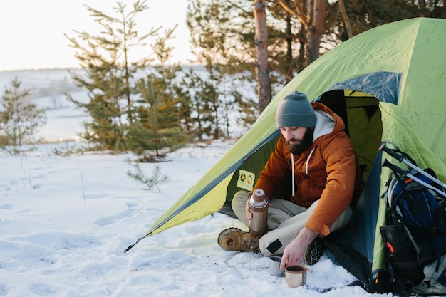 ひげを生やした若い男性がテントの近くの冬の山で休む 帽子にひげを生やし、暖かいジャケットを着た男旅行者が、ハイキングの後に熱いお茶やコーヒーを飲んで体を温める 旅行のライフスタイル