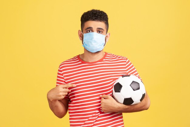 Young bearded man in safety mask and red t-shirt pointing finger at soccer ball on his hand with sad expression, coronavirus quarantine. Indoor studio shot isolated on yellow background