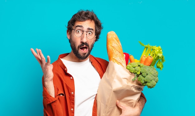 Young bearded man screaming with hands up in the air, feeling furious, frustrated, stressed and upset and holding a vegetables bag