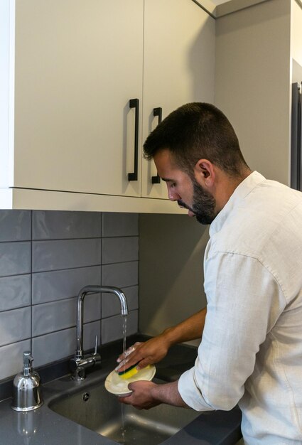 若いあごひげを生やした男が台所の流しで食器を石鹸と水でこすり洗いする