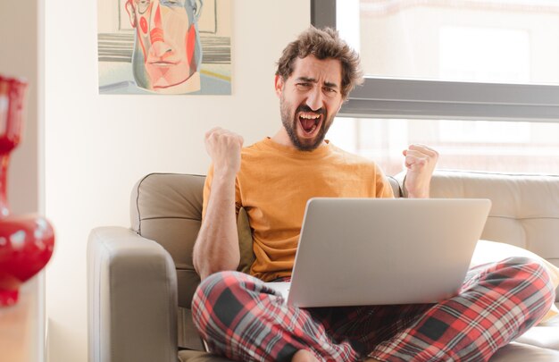 Photo young bearded man shouting aggressively with an angry expression or with fists clenched celebrating success and sitting with a laptop