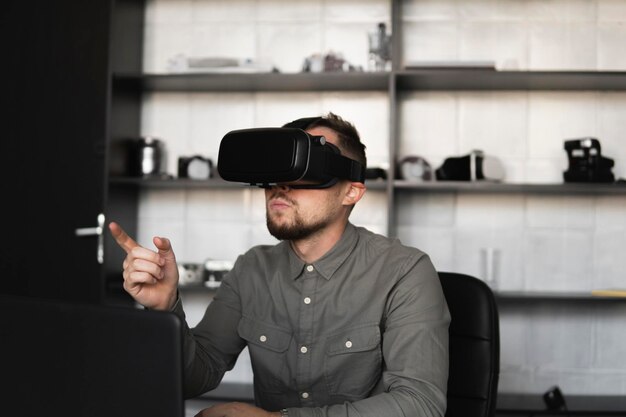 young bearded man sitting against computer with virtual reality glasses trying to touch something