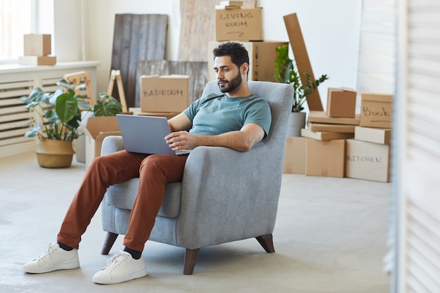 Young bearded man sitting on armchair and using laptop computer he moving to new apartment