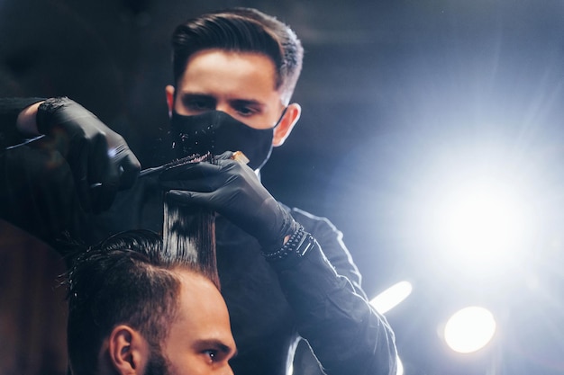 Young bearded man sitting and getting haircut in barber shop by guy in black protective mask