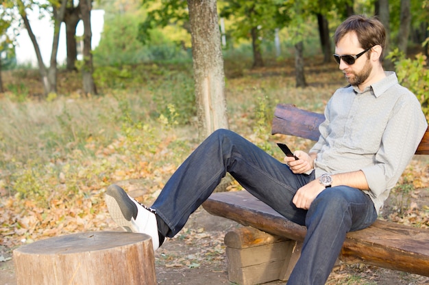 Young bearded man sitting on a wooden bench in the countryside texting on his cellphone or mobile phone