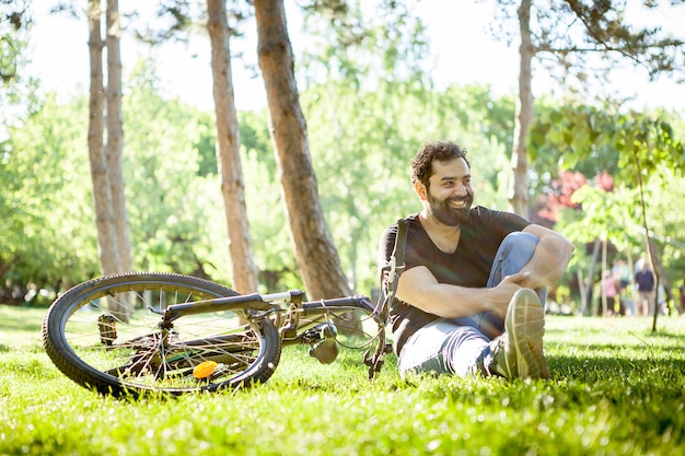 Young bearded man smiles to the camera sitting on the ground in the grass in the park. His bicycle is next to him on the ground