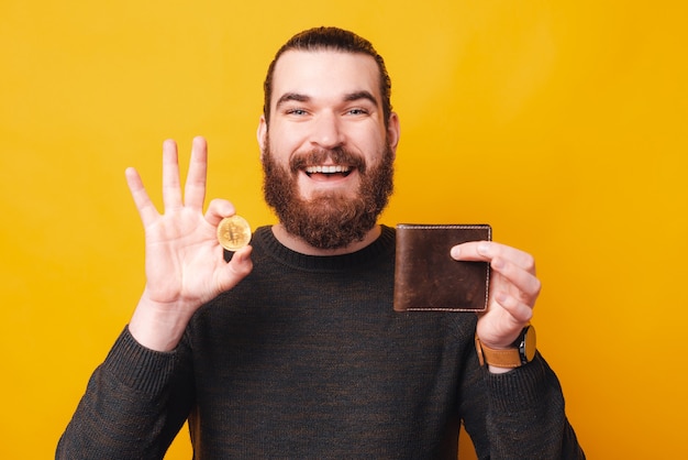 A young bearded man smiling is holding a bitcoin and his wallet looking 