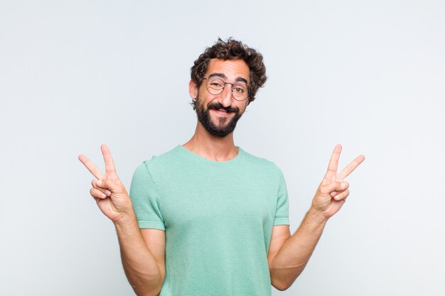 Giovane uomo barbuto sorridente e guardando felice, amichevole e soddisfatto, gesticolando vittoria o pace con entrambe le mani