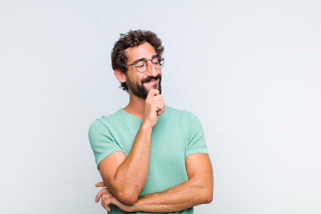 Young bearded man smiling with a happy, confident expression with hand on chin, wondering and looking to the side