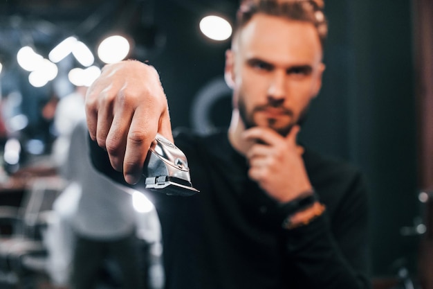 Young bearded man standing in barber shop and holding clipper