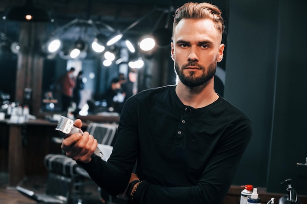 Young bearded man standing in barber shop and holding clipper