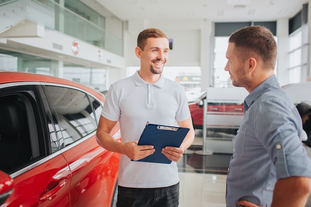 Young bearded man stands in front of customer and smiles. He holds plastic tablet with both hands. People are in front of red and beautfiul car. Customer is serious.