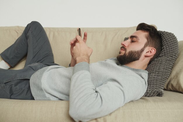Молодой бородатый мужчина в спортивных штанах и толстовке расслабляется на диване в гостиной и прокручивает смартфон во время самоизоляции