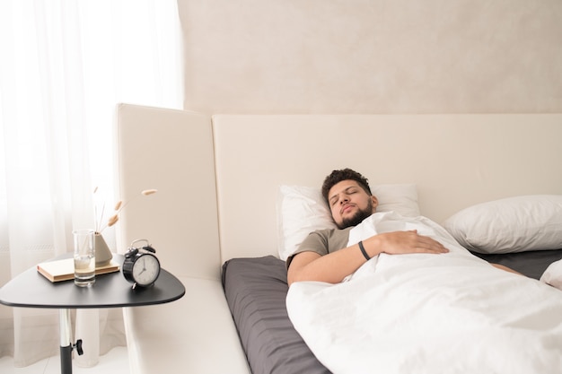 Young bearded man in t-shirt sleeping in large comfortable\
double bed in front of small table with alarm clock, glass of\
water, vase and book