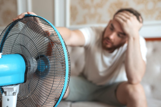 Young bearded man using electric fan at home sitting on couch cooling off during hot weather