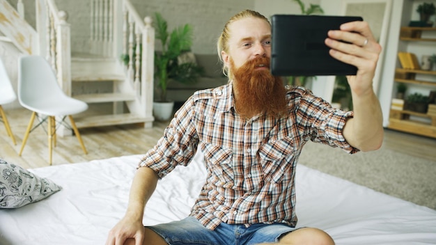 自宅のベッドに座ってビデオチャットを持っているタブレットコンピュータを使用して若いひげを生やした男