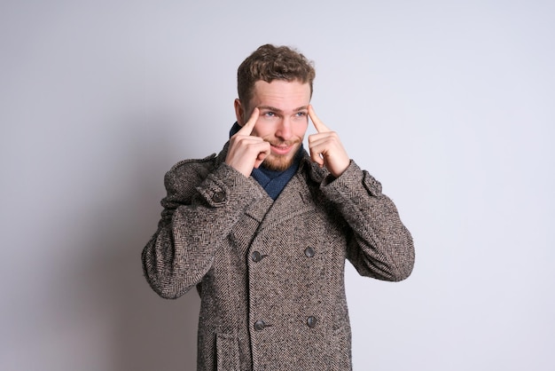 A young bearded man in a warm coat poses emotionally against a light wall