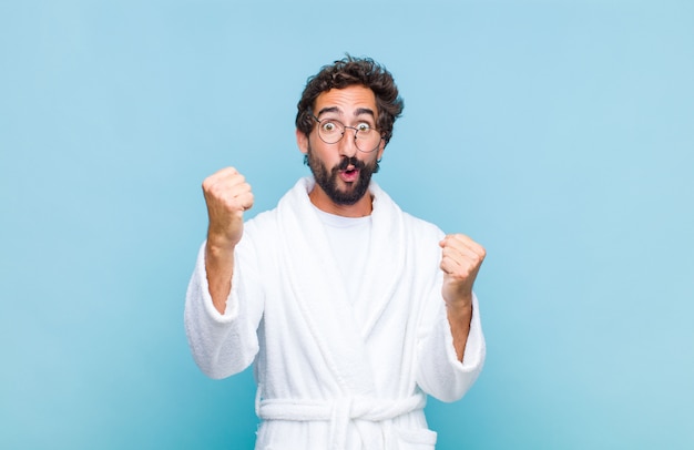 Young bearded man wearing a bath robe celebrating an unbelievable success like a winner, looking excited and happy saying take that!