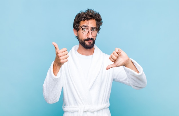 Young bearded man wearing a bath robe feeling confused, clueless and unsure, weighting the good and bad in different options or choices