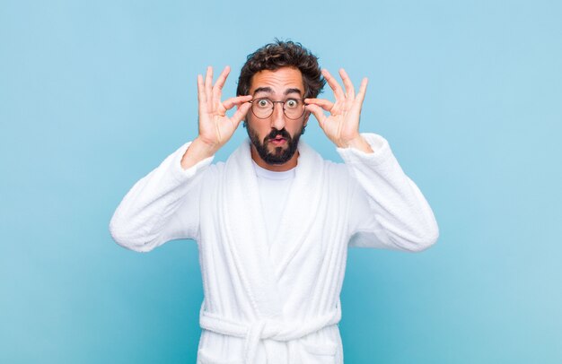 Young bearded man wearing a bath robe feeling shocked, amazed and surprised, holding glasses with astonished, disbelieving look