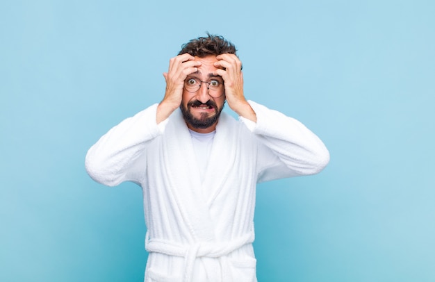 Young bearded man wearing a bath robe feeling stressed and anxious, depressed and frustrated with a headache, raising both hands to head