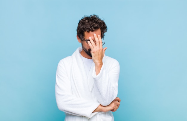 Young bearded man wearing a bath robe looking stressed, ashamed or upset, with a headache, covering face with hand