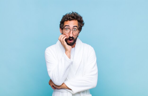 Young bearded man wearing a bath robe open-mouthed in shock and disbelief, with hand on cheek and arm crossed, feeling stupefied and amazed