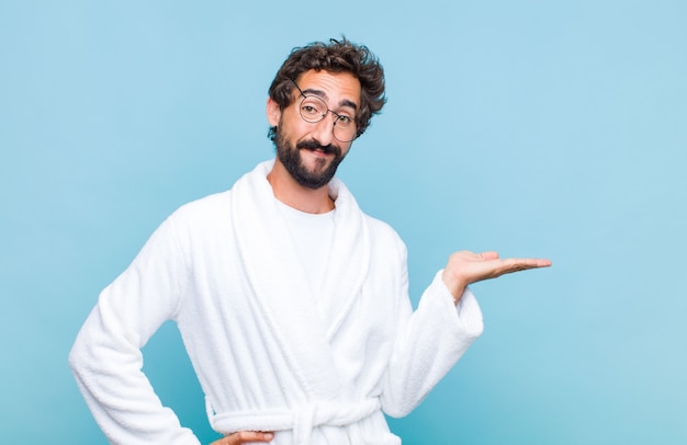 Young bearded man wearing a bath robe smiling, feeling confident, successful and happy, showing concept or idea on copy space on the side