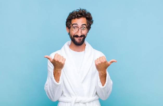 Young bearded man wearing a bath robe smiling joyfully and looking happy, feeling carefree and positive with both thumbs up