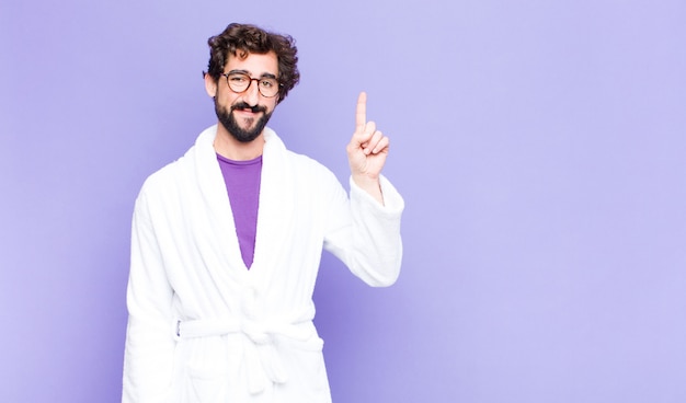 Young bearded man wearing bathrobe smiling cheerfully and happily, pointing upwards with one hand to copy space