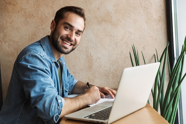 Foto giovane uomo barbuto che indossa una camicia di jeans che scrive con il computer portatile mentre si lavora in un bar al chiuso