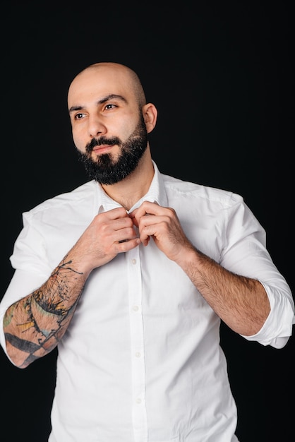 A young bearded man in a white shirt and tattoos stands on a black wall.