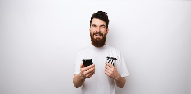 Young bearded man in white shirt on white background holding his phone and a cup of coffee or tea take away or to go.