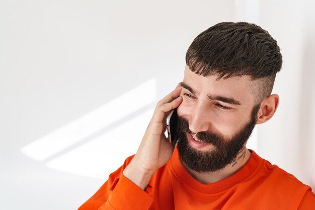 молодой бородатый мужчина с украшениями для носа в оранжевой рубашке разговаривает по смартфону, стоя у белой стены в помещении