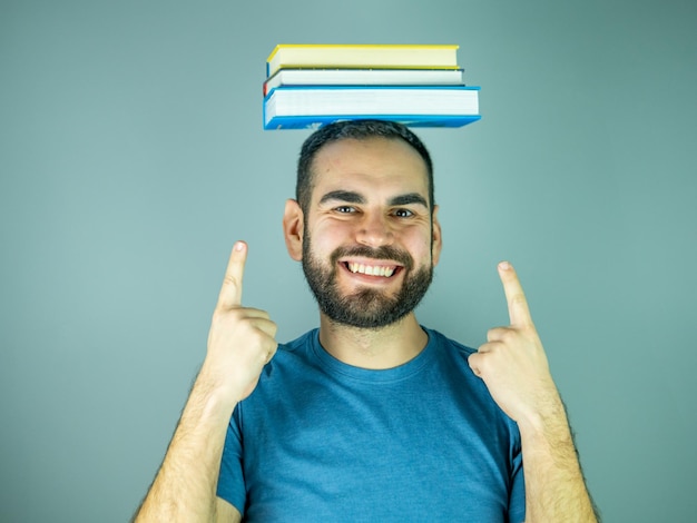 young bearded man with a pile of books on his head pointing up while looking at camera