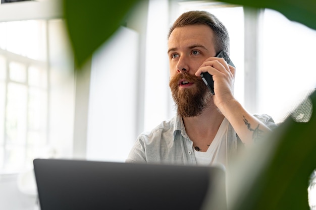 Young bearded man working from home with laptop while talking on the phone