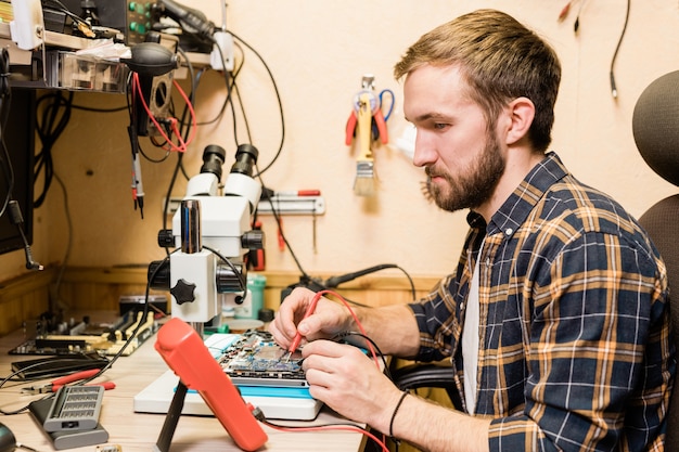 Young bearded mechanic with two soldering-irons sitting by workplace in workshop while repairing broken gadget
